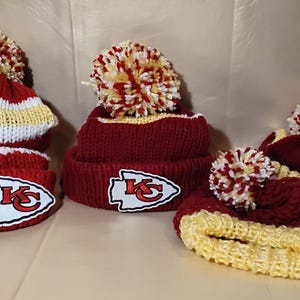 May include: A collection of knitted winter hats in red, white, and yellow. Each hat features a pom-pom and a team logo patch. The hats are designed for cold weather and are likely for sports fans.