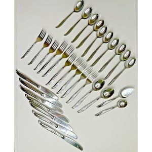 May include: A collection of stainless steel silverware, including spoons, forks, knives, and serving spoons, arranged on a white surface. The silverware has a shiny, reflective finish, and the arrangement suggests a formal dining setting.