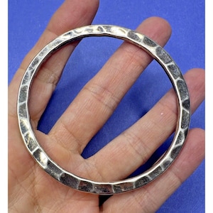 May include: A circular silver-toned bangle bracelet featuring a hammered, textured surface with a reflective metallic finish. The bangle has a consistent rounded profile and a polished appearance, showcasing indentations across the entire outer band.