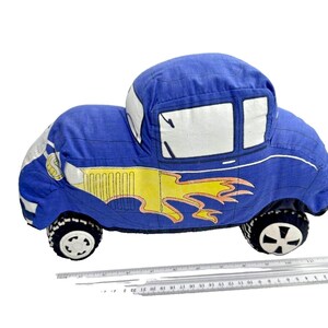 May include: A blue fabric car-shaped pillow featuring a yellow flame graphic on the side, white windows, and black and white wheels. A clear plastic ruler is positioned underneath the pillow, showing a length of approximately 9 inches.