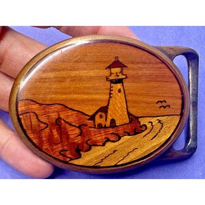 May include: Oval wooden belt buckle featuring a lighthouse scene. The buckle has a brown wooden background with a lighthouse, waves, and birds. The metal frame is a silver color.