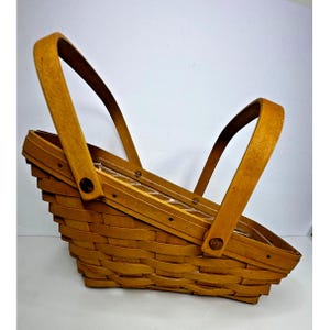 May include: A light brown woven wooden basket with a slanted top profile and two tall, arched handles attached to the sides with dark circular rivets. The basket features a traditional horizontal weave pattern and a sturdy, flat base structure.