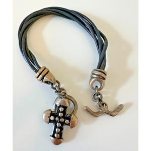 May include: A multi-strand dark green leather bracelet featuring a metallic cross pendant with a black center and raised silver-toned dots. The bracelet includes silver-toned metal end caps and a T-bar toggle clasp closure mechanism.