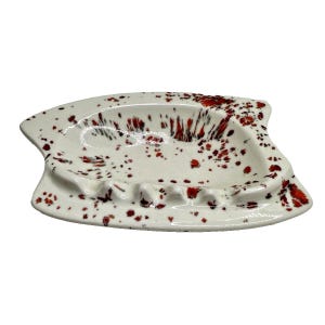 May include: A white ceramic ashtray with an irregular, asymmetrical shape featuring a speckled pattern of red and black paint splatters across the surface. The dish has a shallow basin and molded indentations along the rim for resting items.