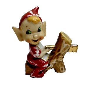 May include: A ceramic figurine of a smiling elf with pointed ears, wearing a red hat and red outfit with white accents. The elf is sitting on a brown tree stump and holding a gold-coloured saw, appearing to be in the process of cutting the wood.