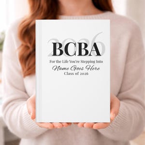 Op de afbeelding: Een wit notitieboek met de tekst "BCBA" in grote, vette letters. Daaronder staat de tekst "For the Life You're Stepping Into Name Goes Here Class of 2026". Het notitieboek wordt voor een persoon gehouden.