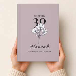 May include: A hardback book with a dusty mauve cover featuring the text "CHAPTER 30" in black, a white floral illustration with dark outlines, the name "Hannah" in a script font, and the phrase "Blooming In Your Own Time" in black text.