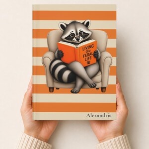 May include: A hardcover book with a striped orange and cream cover featuring an illustration of a raccoon wearing glasses, sitting in a chair, and reading an orange book titled Living the Feral Life. The name Alexandria is printed at the bottom.