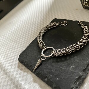 May include: A silver chain bracelet featuring a ring and a pointed spike charm. The bracelet is crafted from interlocking metal rings.