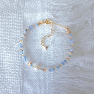 May include: Delicate beaded bracelet featuring a mix of light blue, gold, and cream-colored beads. The bracelet includes a central freshwater pearl and a gold-toned clasp. The bracelet is displayed on a white, textured surface.
