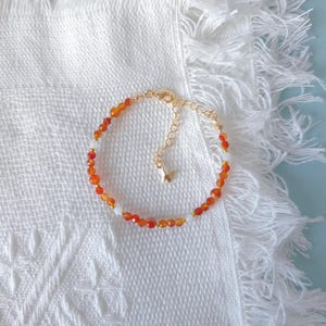 Peut inclure: Un bracelet délicat orné de petites perles de pierres précieuses orange et blanches. Le bracelet est doté d'une chaîne et d'un fermoir dorés. Les perles sont enfilées, créant un design simple mais élégant. Le bracelet est présenté sur une surface blanche texturée.