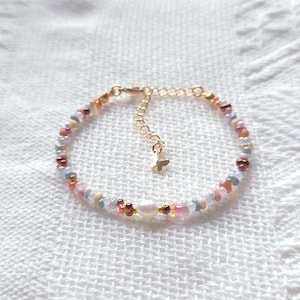 Peut inclure: Un bracelet de perles composé d'une perle d'eau douce blanche centrale entourée de petites perles en verre multicolores dans des tons rose, bleu et doré. Le bracelet comprend une chaînette d'extension en métal doré avec un petit papillon.