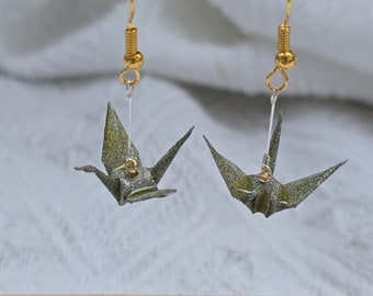May include: A pair of gold dangle earrings with green origami cranes hanging from them. The cranes have a glittery finish.