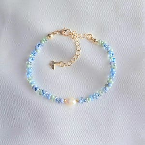 May include: A beaded bracelet featuring a central cream-coloured pearl bead flanked by small light blue and pale green glass beads. The bracelet includes a gold-toned metal lobster clasp, an adjustable chain extension, and a small cross-shaped charm.