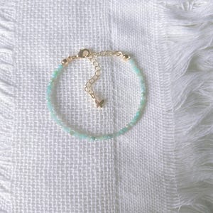 Könnte beinhalten: Zartes Armband mit kleinen, hellblauen Perlen. Das Armband hat eine goldfarbene Kette und einen Verschluss mit einem kleinen Schmetterlingsanhänger. Der Schmuck wird auf einer weißen, strukturierten Oberfläche präsentiert.