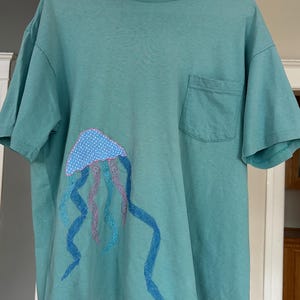 Op de afbeelding: Tealkleurig T-shirt met een zak en een kwalontwerp. De kwal heeft een blauwe stippen kop en vloeiende blauwe en roze tentakels. Het shirt hangt aan een witte hanger.