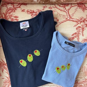 Puede incluir: Dos camisetas de cuello redondo, una azul marino y otra celeste, cada una adornada con un diseño de aceitunas bordadas. Las aceitunas son verdes con lunares blancos y un corazón rojo. La camiseta azul marino tiene una etiqueta "Trash Mail Studios".