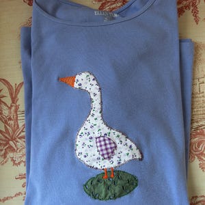 May include: A periwinkle blue t-shirt featuring a goose applique. The goose is white with a purple floral pattern and a purple checked heart. It has an orange beak and feet, and sits on a green leaf.