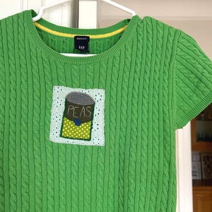 Könnte beinhalten: Ein grüner Kurzarmpullover mit Zopfmuster. Der Pullover hat einen Aufnäher mit einer Illustration einer Erbsendose. Das Wort "PEAS" ist auf dem Aufnäher aufgedruckt. Der Ausschnitt ist gelb eingefasst.