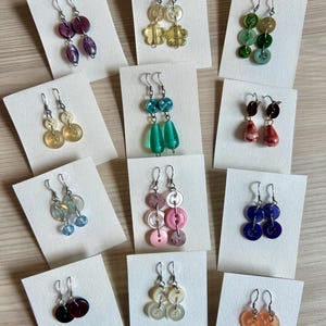 Könnte beinhalten: Eine Sammlung handgefertigter Ohrringe, präsentiert auf weißen Karten. Die Ohrringe bestehen aus Knöpfen in verschiedenen Farben wie Lila, Gelb, Grün, Blau und Rosa. Jedes Paar hat einen silbernen Haken.