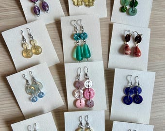 May include: A collection of handmade earrings displayed on white cards. The earrings feature various button designs in colors like purple, yellow, green, blue, and pink. Each pair has a silver hook.