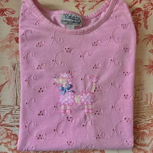 Pode incluir: Uma camiseta rosa dobrada com gola redonda e detalhes vazados. Apresenta um poodle bordado com laço azul e bolinhas. A etiqueta diz "Tradition Country Collection".