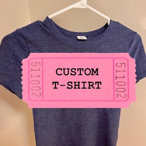 May include: A navy blue short-sleeved t-shirt on a white hanger. A pink ticket-shaped label is overlaid on the shirt, with the words "CUSTOM T-SHIRT" printed in black.
