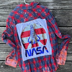 May include: Red and blue checked flannel shirt with a white patch on the back. The patch features a sequined rocket ship and the word "NASA" in blue sequins.