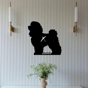 May include: A black poodle dog silhouette with the name "MASIE" beneath it, mounted on a white, vertically-lined wall. Two gold and white sconces flank the dog. A vase of flowers is positioned below the dog.
