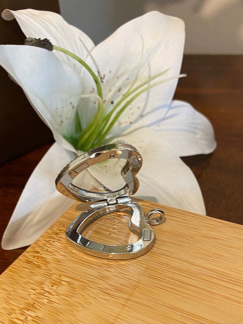 Puede incluir: Un medall&oacute;n de plata en forma de coraz&oacute;n con una ventana de vidrio transparente. El medall&oacute;n est&aacute; abierto y el interior es visible. El medall&oacute;n est&aacute; sobre una superficie de madera.