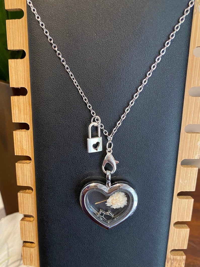 Puede incluir: Un collar de cadena plateada con un dije en forma de coraz&oacute;n. El dije tiene una ventana de vidrio transparente que revela una flor seca en su interior. Un peque&ntilde;o dije de candado plateado cuelga de la cadena.