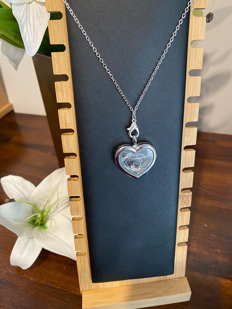 Puede incluir: Un collar de plata con un dije en forma de coraz&oacute;n y un inserto de vidrio transparente. El dije est&aacute; colgado en un expositor de madera con ranuras para joyas.