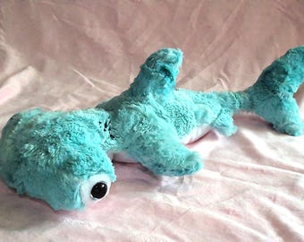 May include: A plush blue hammerhead shark toy with large, black eyes. The toy is lying on a pink surface.