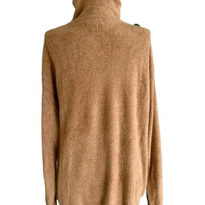 May include: A tan-coloured turtleneck jumper with long sleeves. The jumper has a soft, textured appearance and a relaxed fit. The turtleneck is folded over. The jumper is made of a warm, comfortable material.