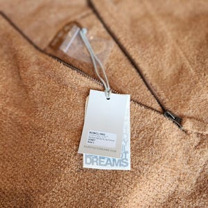 May include: A close-up of a tan-coloured, textured pullover sweater with a zipper and a tag. The tag reads "Barefoot Dreams" and includes the text "COZYCHIC LITE" and "HONEY". The sweater has a soft, plush appearance.