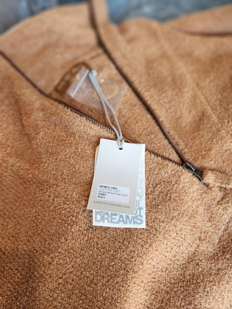 May include: A close-up of a tan-coloured, textured pullover sweater with a zipper and a tag. The tag reads "Barefoot Dreams" and includes the text "COZYCHIC LITE" and "HONEY". The sweater has a soft, plush appearance.