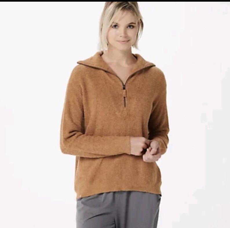 May include: A tan-coloured, long-sleeved pullover sweater with a zip closure at the neckline. The sweater has a collared neckline and a soft, textured appearance. The model is wearing grey trousers.