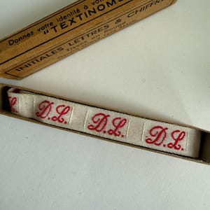 May include: A beige fabric ribbon featuring the repeating red embroidered initials D.L. inside a brown cardboard box. The box lid displays the text Donnez votre identit&eacute; &agrave; votre linge avec les TEXTINOMS, INITIALES LETTRES & CHIFFRES.