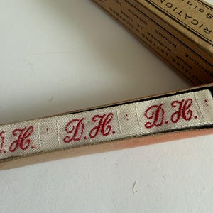 May include: A beige fabric ribbon featuring the embroidered red initials D.H. repeated along its length, housed in a vintage cardboard box with French text including the words FABRICATION FRAN&Ccedil;AISE and SAINT-&Eacute;TIENNE printed on the lid.