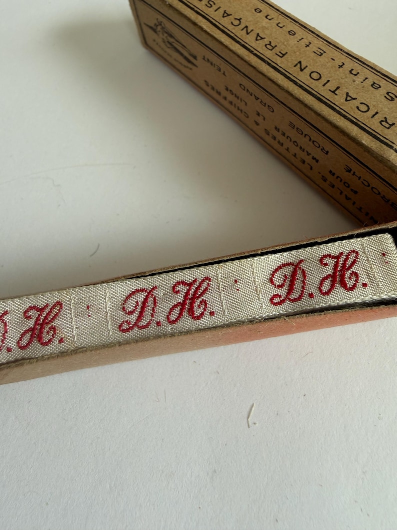 May include: A beige fabric ribbon featuring the embroidered red initials D.H. repeated along its length, housed in a vintage cardboard box with French text including the words FABRICATION FRAN&Ccedil;AISE and SAINT-&Eacute;TIENNE printed on the lid.