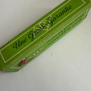 May include: A rectangular green cardboard box with white border lines. The text reads Une Grosse Garantie, INITIALES ET CHIFFRES POUR MARQUER LE LINGE. A small illustration shows a red and black bird-like figure holding a white object.