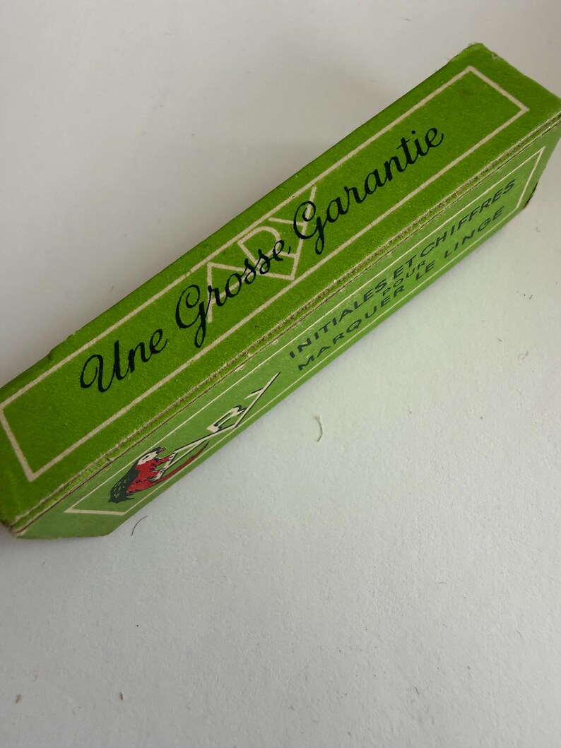 May include: A rectangular green cardboard box with white border lines. The text reads Une Grosse Garantie, INITIALES ET CHIFFRES POUR MARQUER LE LINGE. A small illustration shows a red and black bird-like figure holding a white object.