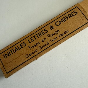 May include: A rectangular tan cardboard label with a black border and black printed text that reads INITIALES LETTRES & CHIFFRES, Tiss&eacute;s en Rouge, and Garanti Grand Teint Absolu. The label is oriented diagonally across the frame.