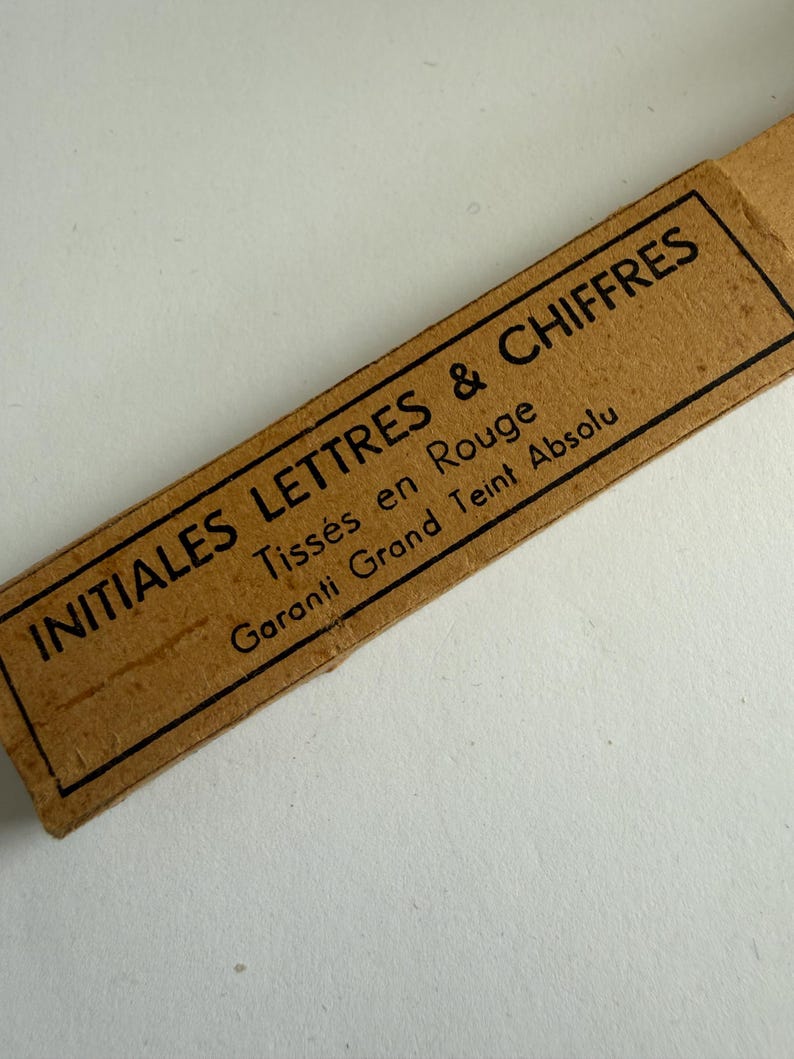 May include: A rectangular tan cardboard label with a black border and black printed text that reads INITIALES LETTRES & CHIFFRES, Tiss&eacute;s en Rouge, and Garanti Grand Teint Absolu. The label is oriented diagonally across the frame.