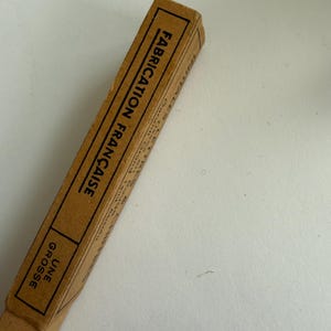 May include: A rectangular tan cardboard box with black text printed on the side. The text reads FABRICATION FRAN&Ccedil;AISE and UNE GROSSE, enclosed within black rectangular borders. The box has a simple, utilitarian design with visible paper edges.