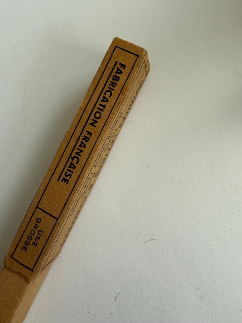 May include: A rectangular tan cardboard box with black text printed on the side. The text reads FABRICATION FRAN&Ccedil;AISE and UNE GROSSE, enclosed within black rectangular borders. The box has a simple, utilitarian design with visible paper edges.