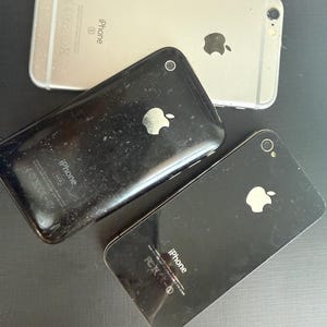 May include: Three smartphones including one silver iPhone 6s and two black iPhones. Each device features the Apple logo on the back, a rear camera lens, and printed text including the word iPhone, regulatory markings, and storage capacity details.