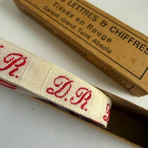 Könnte beinhalten: Eine Rolle aus cremefarbenem Stoffband mit wiederkehrenden, in roter Schreibschrift gestickten Initialen D.R., in einer rechteckigen Pappschachtel mit der Aufschrift INITIALES LETTRES & CHIFFRES Tissés en Rouge Garanti Grand Teint Absolu.