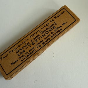 May include: A rectangular tan cardboard box featuring black printed French text that reads: Pour Pensionnats, H&ocirc;tels, Linge de Maison, demandez &agrave; votre fournisseur Les TEXTINOMS, Noms tiss&eacute;s en entier, initiales suivies de chiffres, etc., Livraison 10 jours environ.