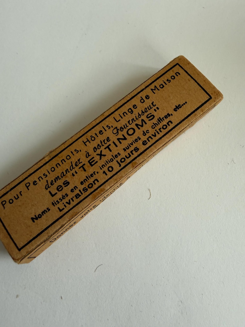 May include: A rectangular tan cardboard box featuring black printed French text that reads: Pour Pensionnats, H&ocirc;tels, Linge de Maison, demandez &agrave; votre fournisseur Les TEXTINOMS, Noms tiss&eacute;s en entier, initiales suivies de chiffres, etc., Livraison 10 jours environ.
